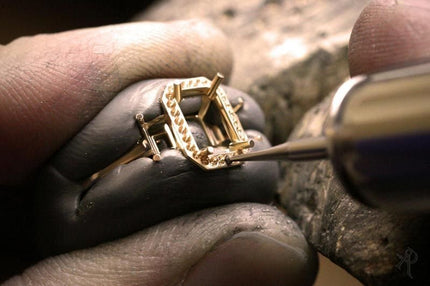 Jeweler's hands crafting a delicate gold ring setting. The jeweler is carefully using a small tool to place tiny prongs around the center stone, which is likely a diamond or other precious gem. The intricate details and precision of the work highlight the artistry and skill involved in creating fine jewelry.